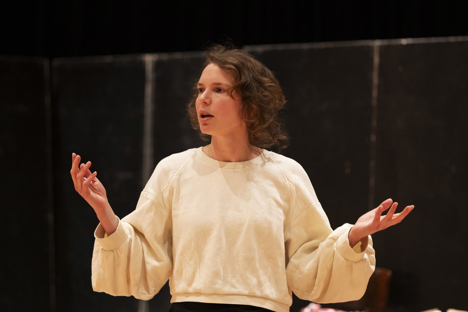 We zien Liliane Brakema tijdens de repetities van Blue Hour. Ze lijkt iets te zeggen en heeft een open houding met haar armen uitgestrekt en haar handpalmen naar boven. 